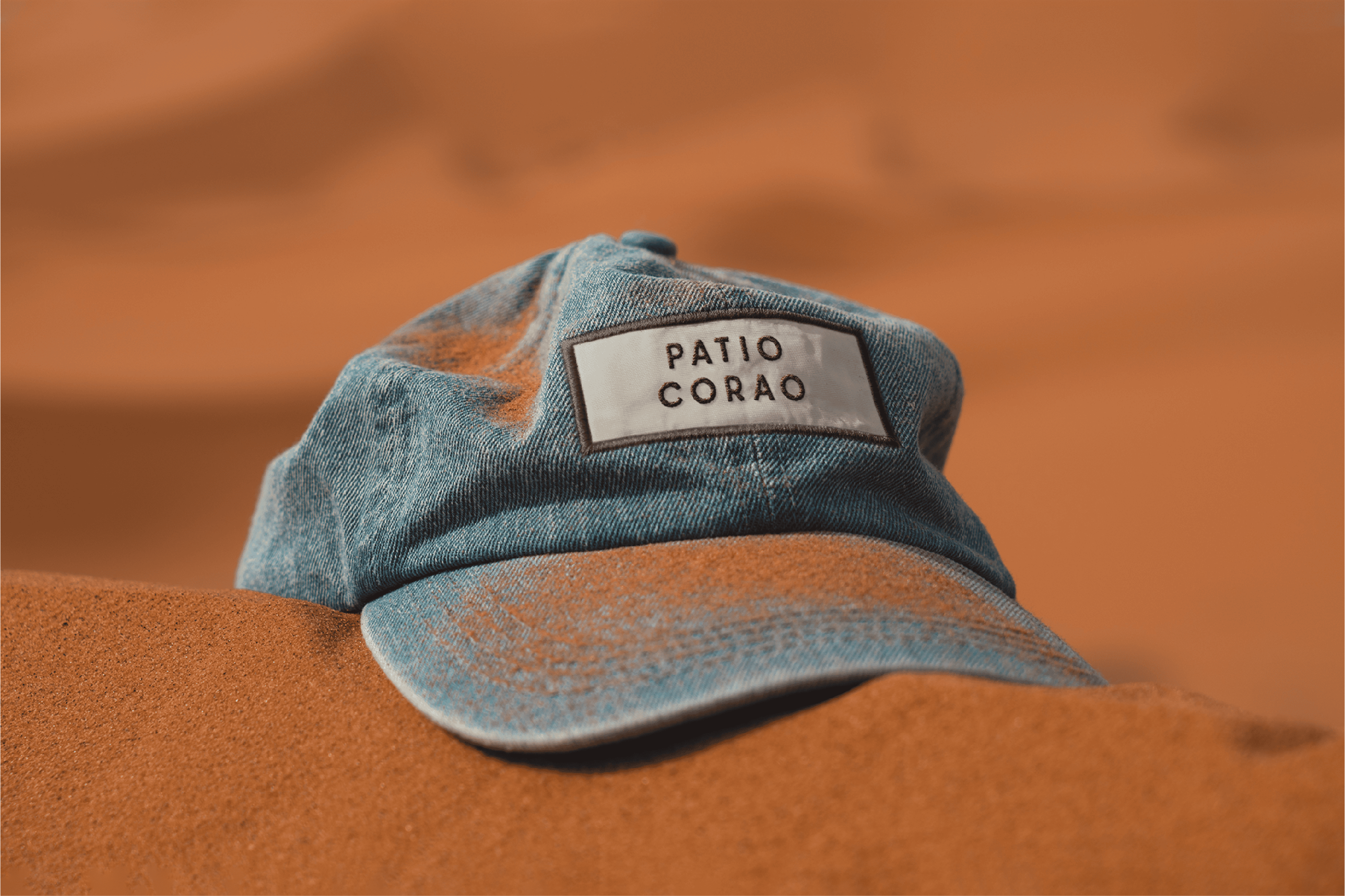 Denim cap with "Patio Corao" label on sandy background, stylish accessory.