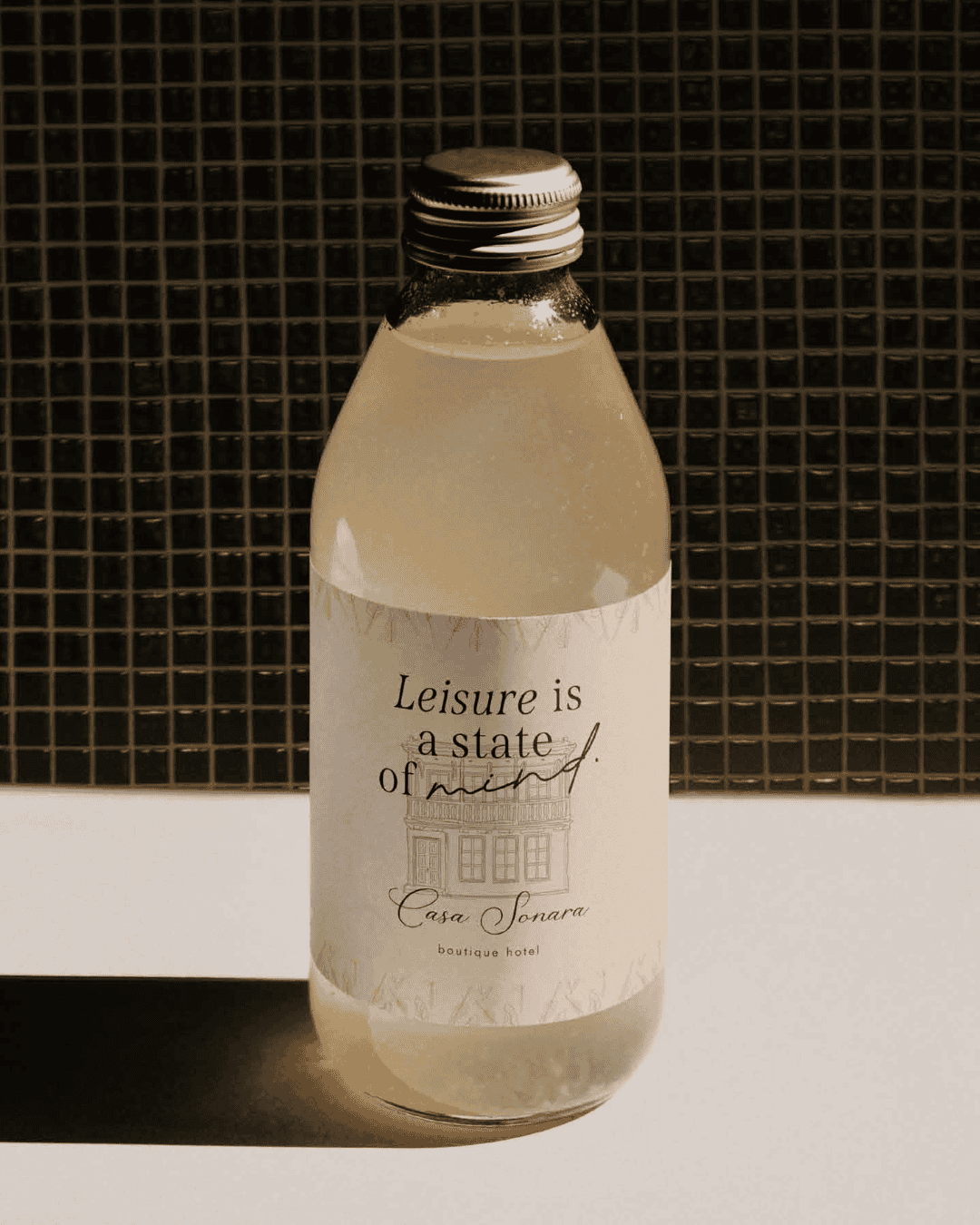 Leisure drink bottle with a silver cap, featuring a minimalist label, placed against a black tiled b.