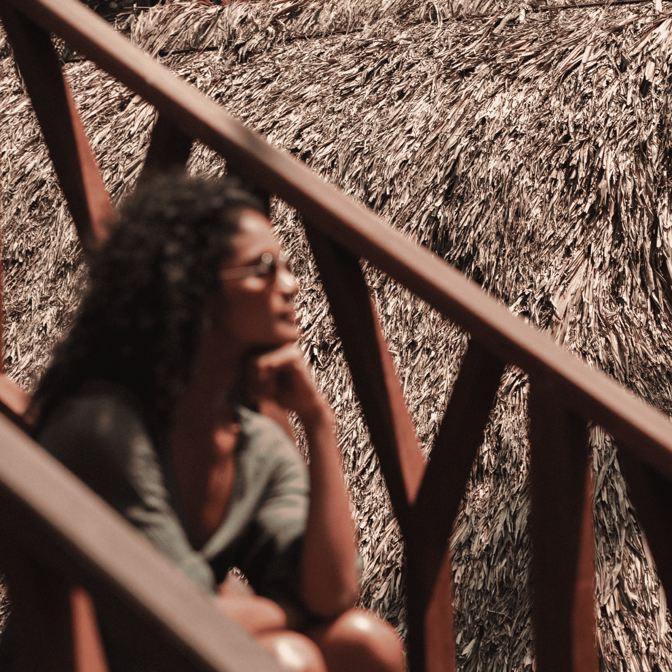 Relaxing woman sitting on a wooden staircase near a thatched roof, enjoying sunny weather and nature.