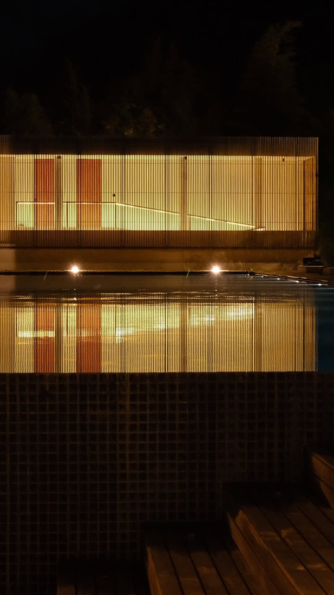 Outdoor swimming pool illuminated by soft lighting with a stylish wooden privacy fence at night.