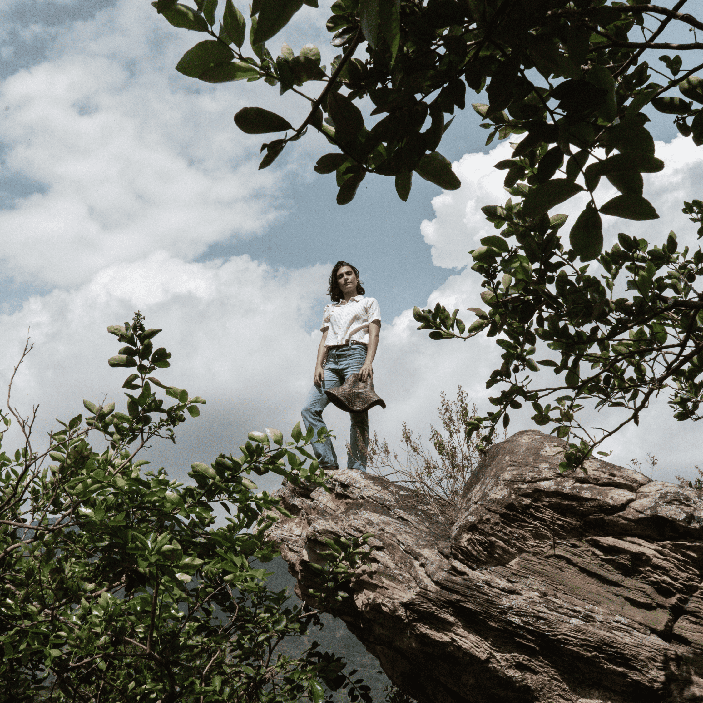 Woman standing on rocky cliff surrounded by greenery and cloudy sky, showcasing outdoor adventure an.