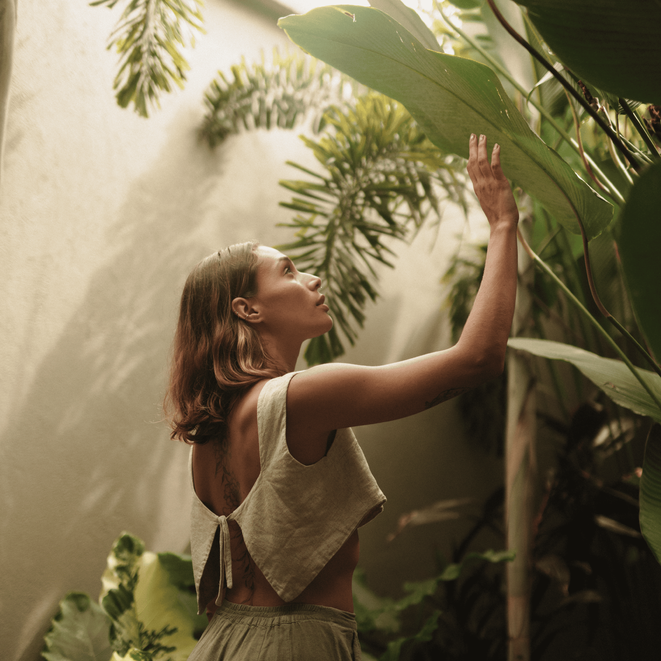 Woman in tropical setting touching large green leaf, lush plants around.