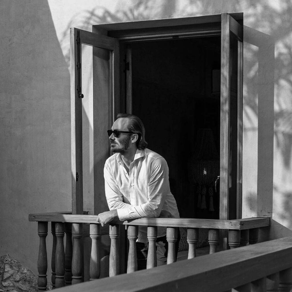 Man with sunglasses leaning on balcony railing near open window.